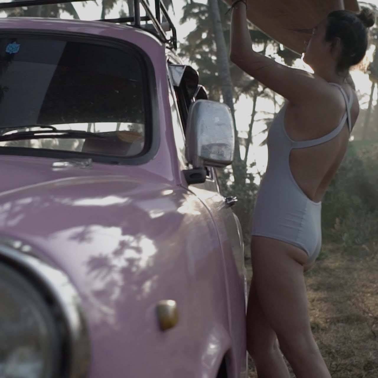 Girl in Coto swimwear lifting surfboard from a classic car at an Indian beach