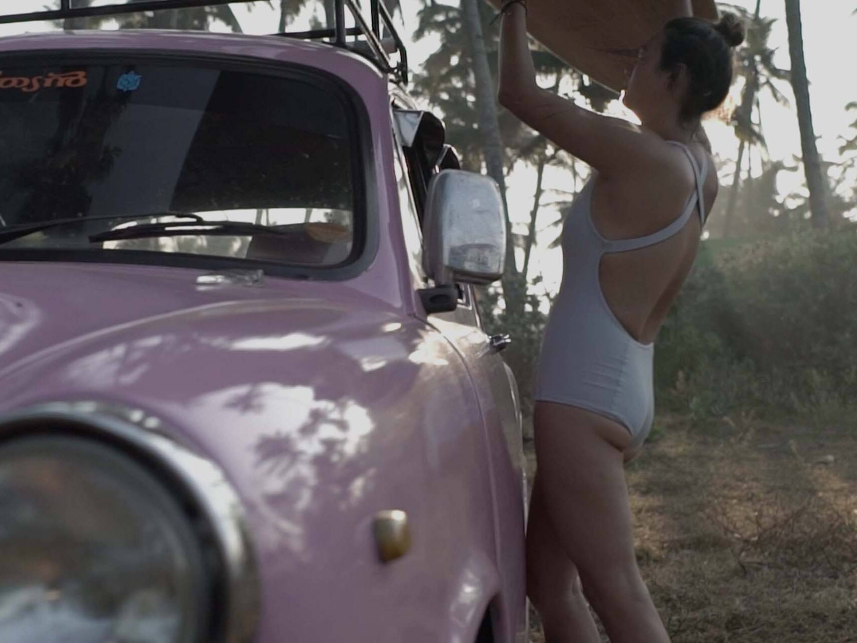 Girl in Coto swimwear lifting surfboard from a classic car at an Indian beach