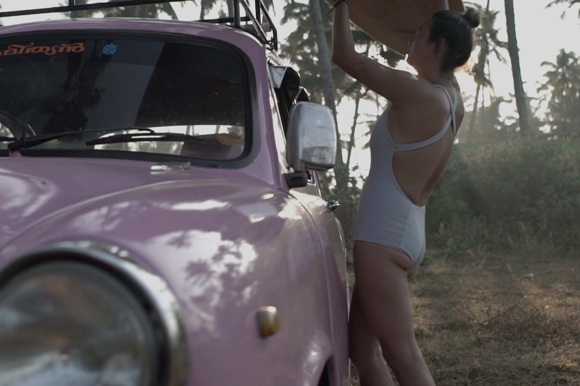 Girl in Coto swimwear lifting surfboard from a classic car at an Indian beach